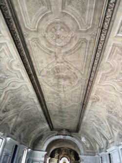 Vista panorâmica do teto em abóbada com estuques cinzentos ao longo da nave da igreja. Ao fundo, o altar com nicho dourado emoldurado por arco de volta perfeita.